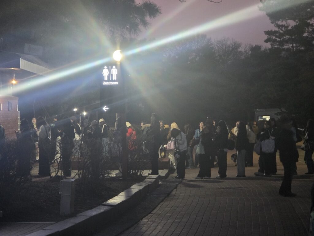 nct-dream-seoul-concert-2026-womens-bathroom-line A documentary-style photograph capturing the overwhelming logistical challenge for female fans at the NCT DREAM Seoul Concert 2026. The image focuses on a seemingly endless queue of women, with hundreds waiting under the brightly lit white canopy of the KSPO DOME, illustrating the "Wall of Patience" required to use the restroom just moments before the show begins.