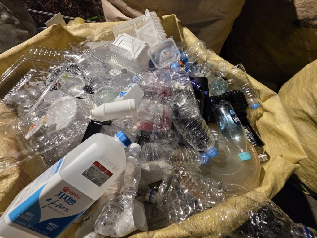 pet-bottle-and-plastic-containers-recycling-korea. A close-up view of a recycling bin filled with clear PET bottles and plastic food containers, showing the high volume of daily waste in Korea.