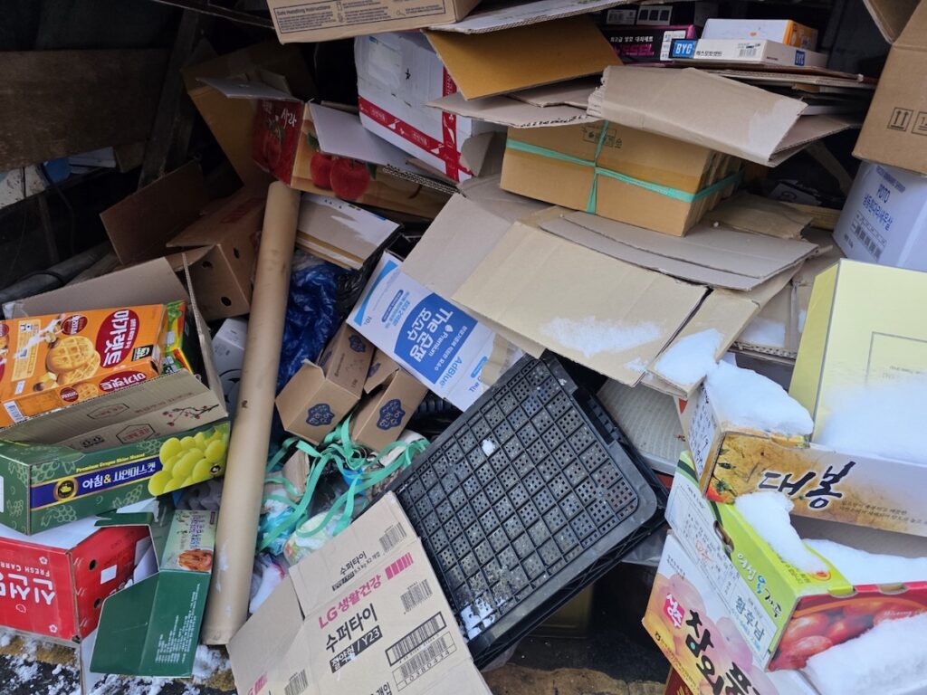 recycling-in-korea-outdoor-paper-collection-winter A pile of flattened cardboard boxes covered in snow at an outdoor collection point, demonstrating the reality of recycling in Korea during the winter season.
