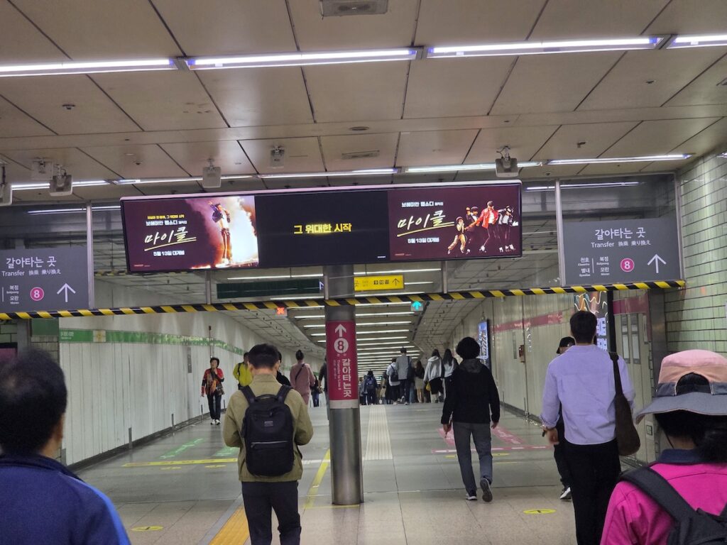 seoul-metro-jamsil-station-transfer-gates-line-8 A perspective view looking down a busy transfer corridor at Jamsil Station. Overhead, a large, widescreen digital display hangs, showing two distinct advertisements. The left advertisement has Korean text 'λ§μ΄ν΄' (Michael) and image of people on stage, while the central black section features the yellow text 'κ·Έ μλν μμ' (The Great Beginning). Below the ads, people are walking towards several automated internal transfer gates that divide the corridor. Signage hanging from the ceiling on both sides clearly displays the pink Line 8 circle with an arrow pointing straight ahead and the text 'κ°μνλ κ³³' (Transfer). Yellow line 2 signage is also visible further in the background. Commuters are walking in both directions.