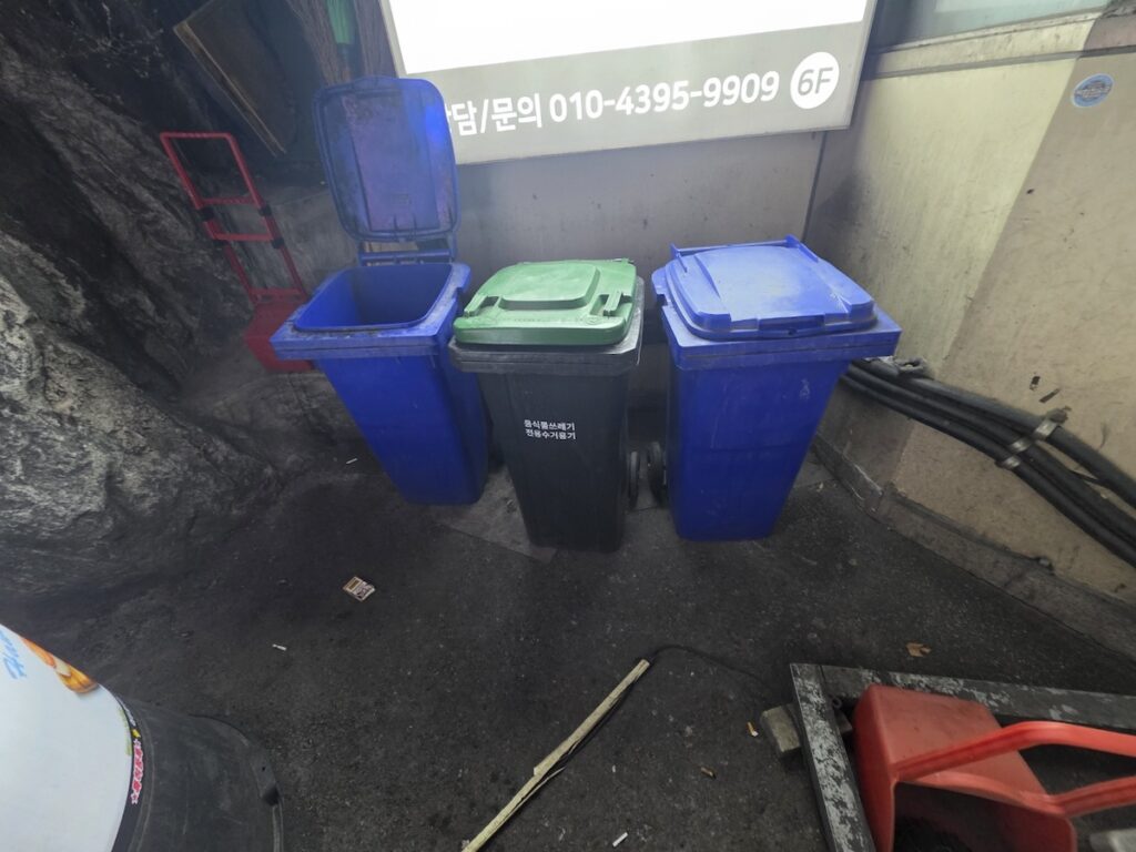 seoul-recycling-system-blue-green-bins Three large waste bins (two blue, one green) positioned under a metal gas pipe at the corner of a stone building.