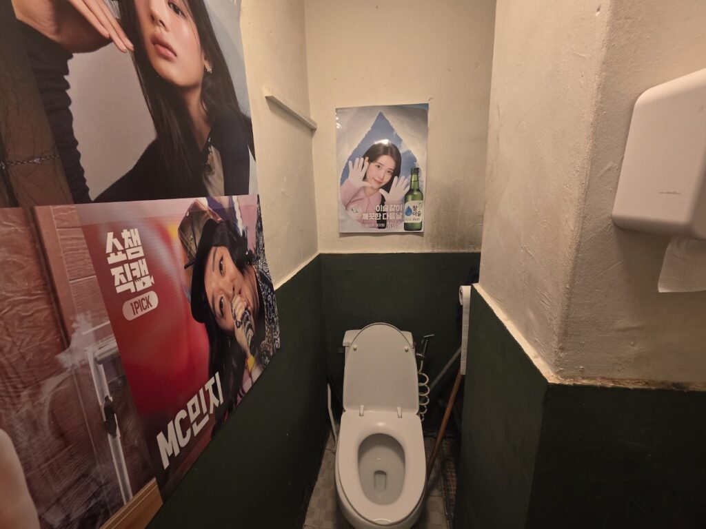 seoul-restroom-rituals-2026-konkuk-pub-aesthetic The aesthetic contrast of K-pop imagery against aged bathroom tiles in a Konkuk University pub, exploring Seoul Restroom Rituals 2026.