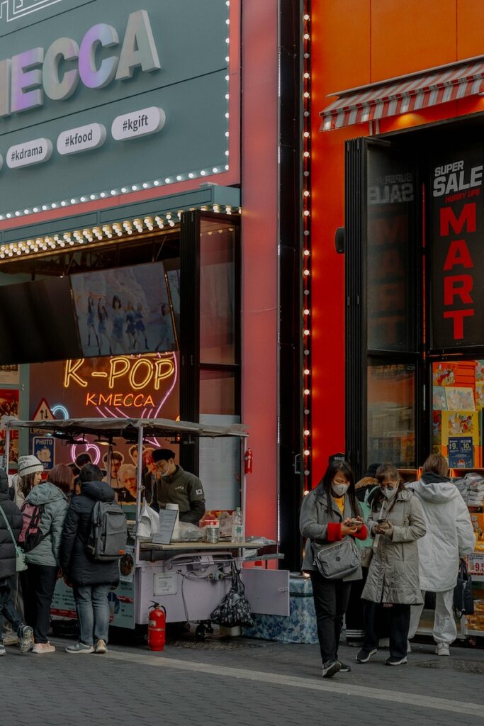 “A lively street in Seoul with K-pop music playing from shops and colorful idol billboards glowing at night, capturing the vibrant pop culture atmosphere of Korea.”