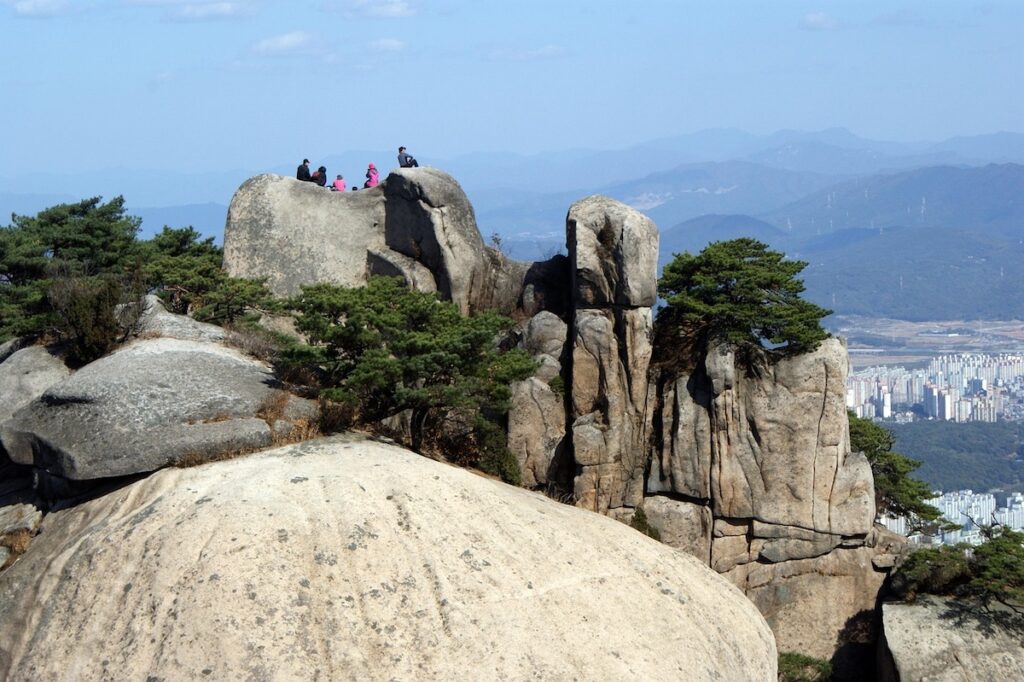 seoul-subway-exit-logic-and-gastronomy-urban-peak Hikers resting on a scenic rock peak overlooking a high-rise city, representing the natural escape reachable by understanding Seoul Subway Exit Logic and Gastronomy.