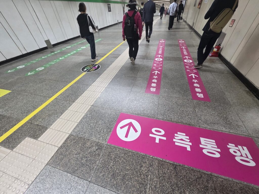 seoul-subway-floor-guide-sticker-line-8-pink-jamsil A closeup photograph taken on the stone tiled floor of a Seoul subway station transfer corridor. Prominently in the foreground is a large, dark pink rectangular floor guide sticker with a white upward arrow. It features the text 'μ°μΈ‘ν΅ν' (Right-hand Traffic) in large white Korean letters. Smaller white text below includes the Line 8 symbol and destination station names: Bokjeong, Moran. Behind it, a continuous line of similar pink guide stickers stretches down the long corridor, leading toward the Line 8 platforms. A single green Line 2 guide sticker is visible on the far left. A few commuters are walking along the right side of the corridor, following the directional flow defined by the pink stickers.
