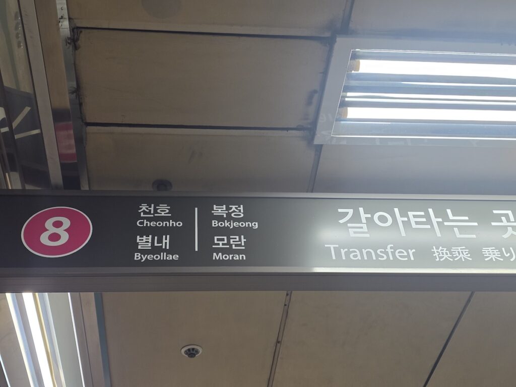 A closeup photograph of a Seoul Subway Line 8 transfer sign hanging from the ceiling. On the left, the dark pink Line 8 circular symbol with the number '8' is clearly visible. Next to it, in two columns, four station names are listed in Korean and English: Cheonho, Byeollae (on the left column), and Bokjeong, Moran (on the right column). On the far right, the large white text '갈아타는 곳' (Transfer) is written in Korean, English (Transfer), Chinese (換乘), and Japanese (乗り換え). The background is a clean white panel under fluorescent lights.