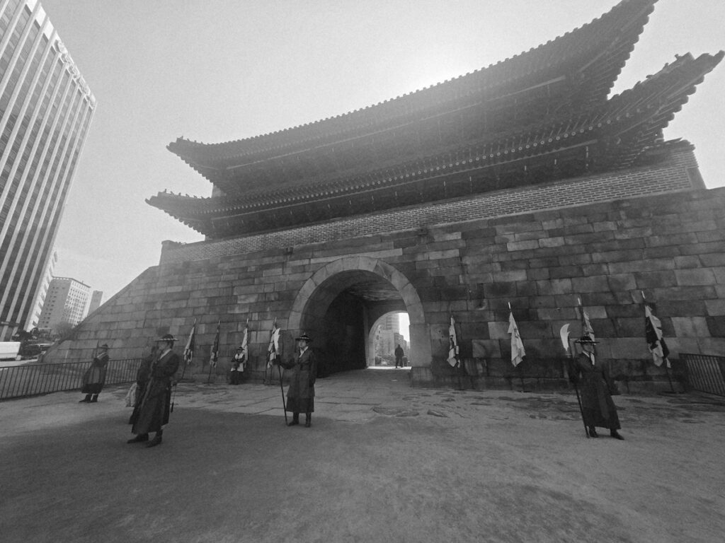 sungnyemun-gate-pasu-ritual-black-and-white-wide-view A black and white wide-angle shot of the Sungnyemun Gate with several guardians stationed in front, capturing the timeless architectural resilience of the fortress.