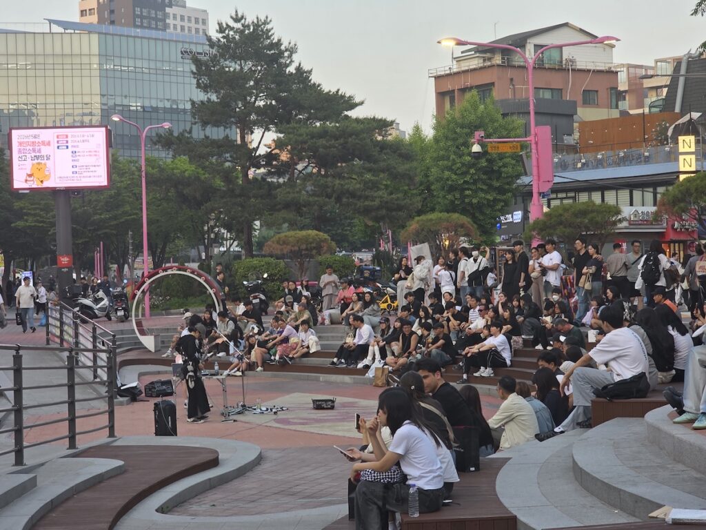 traditional-hongdae-r2-busking-zone-performance A wide-angle view of a tiered public plaza within the Hongdae R2 Busking Zone, showing a solo musician performing at dusk while hundreds of people sit on the steps watching and listening, with large digital screens in the background.