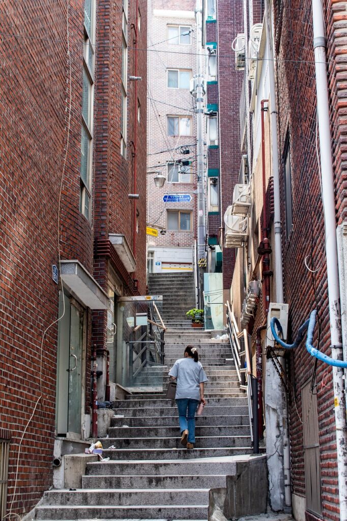 A woman walking up a steep narrow staircase in a traditional red-brick residential neighborhood in Seoul.