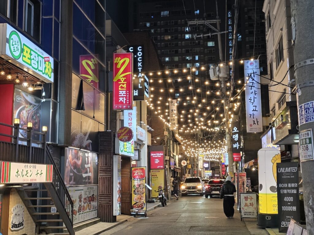 travel-korea-on-a-budget-kondae-nightlife. A vibrant night view of a dining street in Kondae with glowing neon signs, a top destination to travel Korea on a budget.