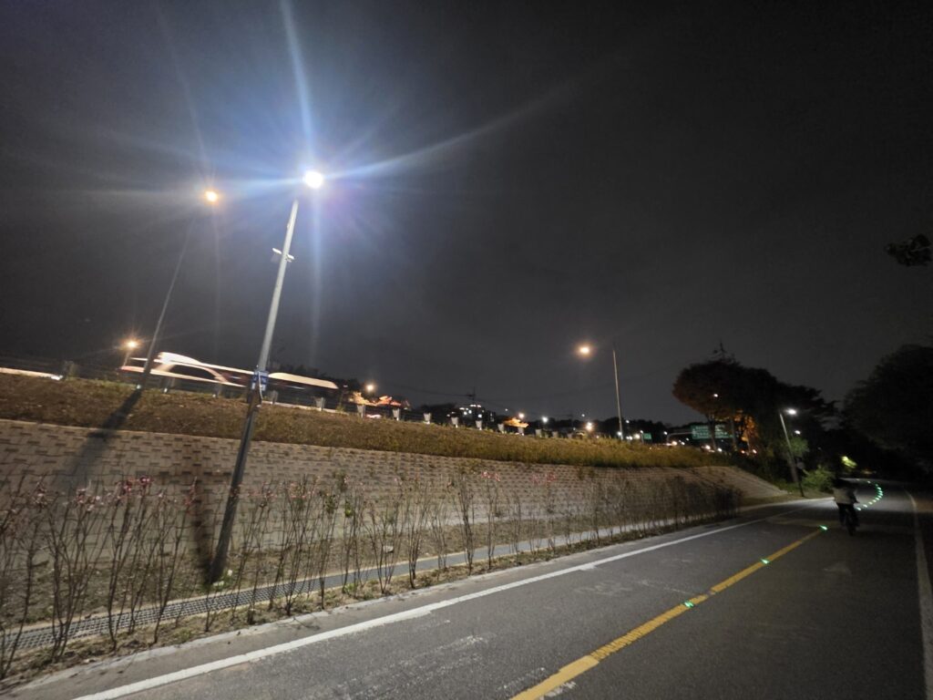 A peaceful night walk along the Han River in Seoul, with the bright traffic of Olympic-daero visible on the right, showing the contrast between calm and motion.