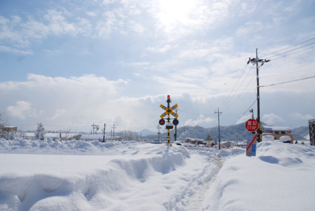 winter-train-korea-snowy-east-sea-railway A scenic view of a train traveling through a heavy snowstorm along the East Coast of South Korea, with tracks and surrounding landscapes covered in deep white snow